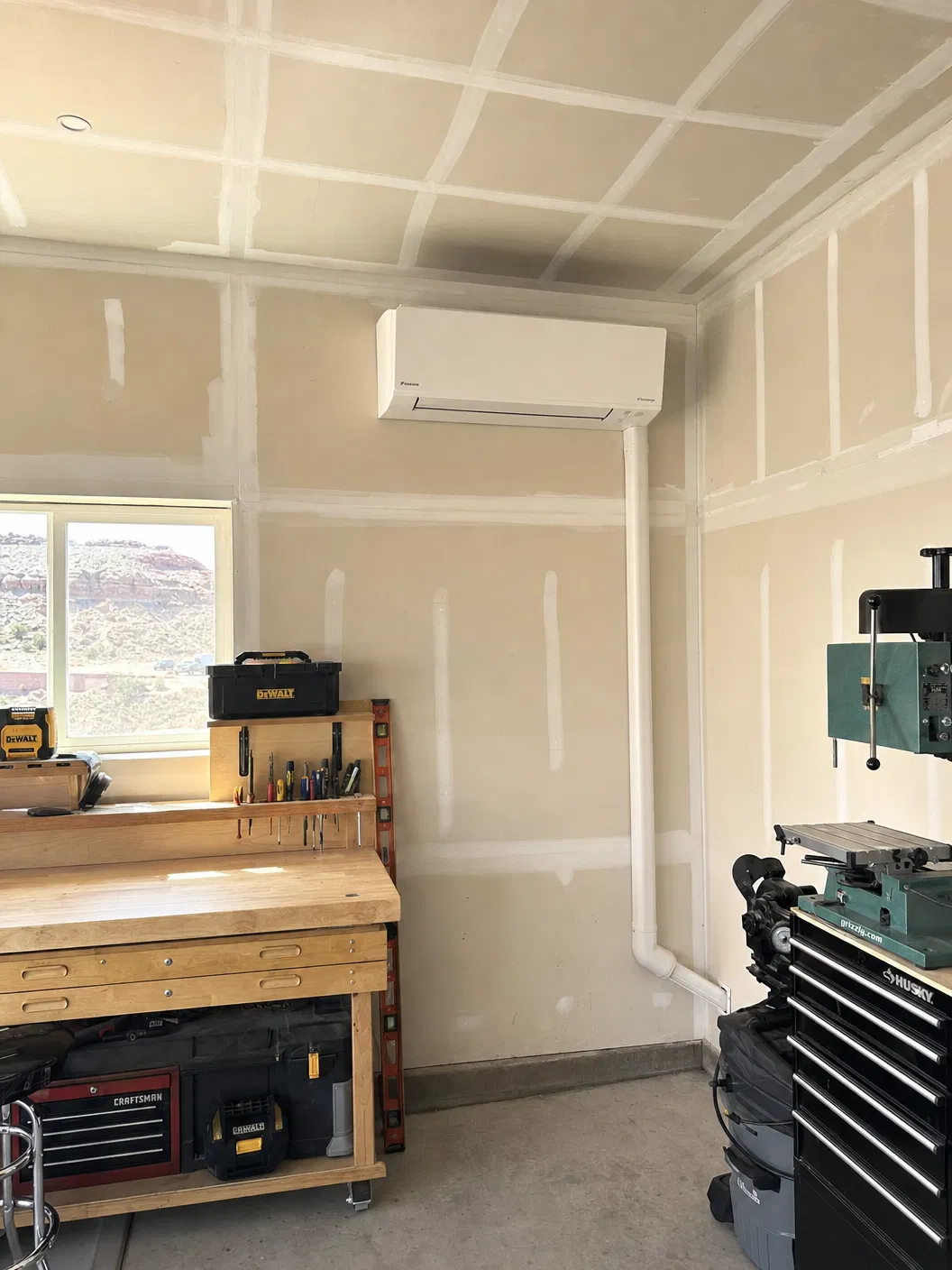 Mini-split indoor head unit installed in a residential garage workshop with workbench and power tools, line set cover running down the wall, Southern Utah red rock visible through window