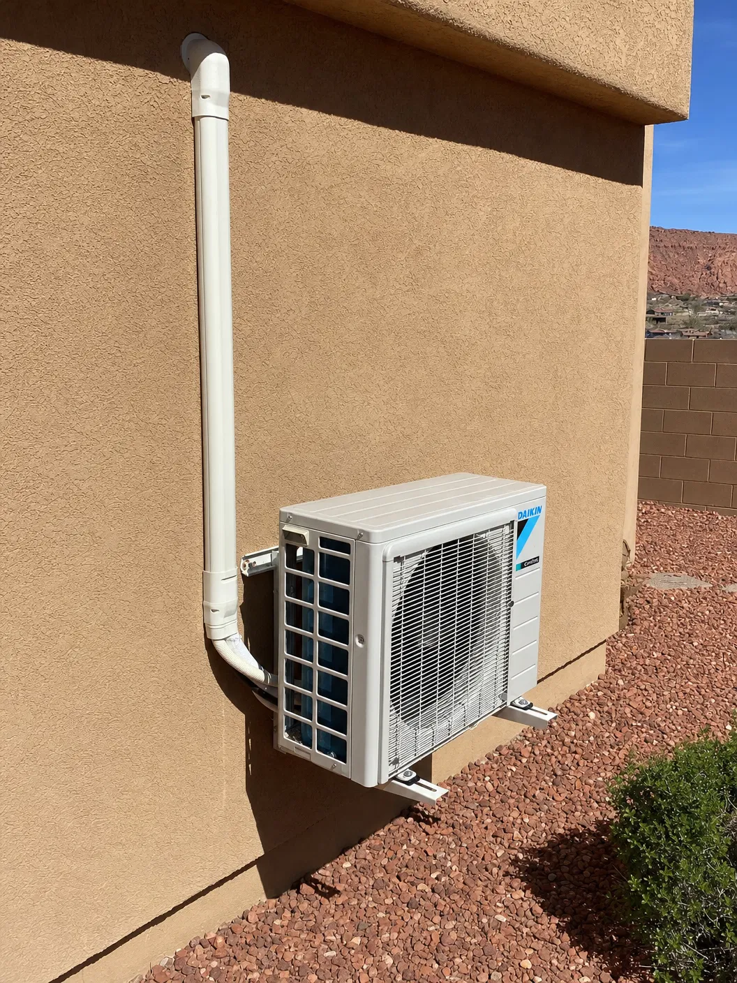 Daikin outdoor mini-split condenser mounted on wall brackets against tan stucco exterior wall with white line set cover and red rock desert landscaping in St. George, Utah